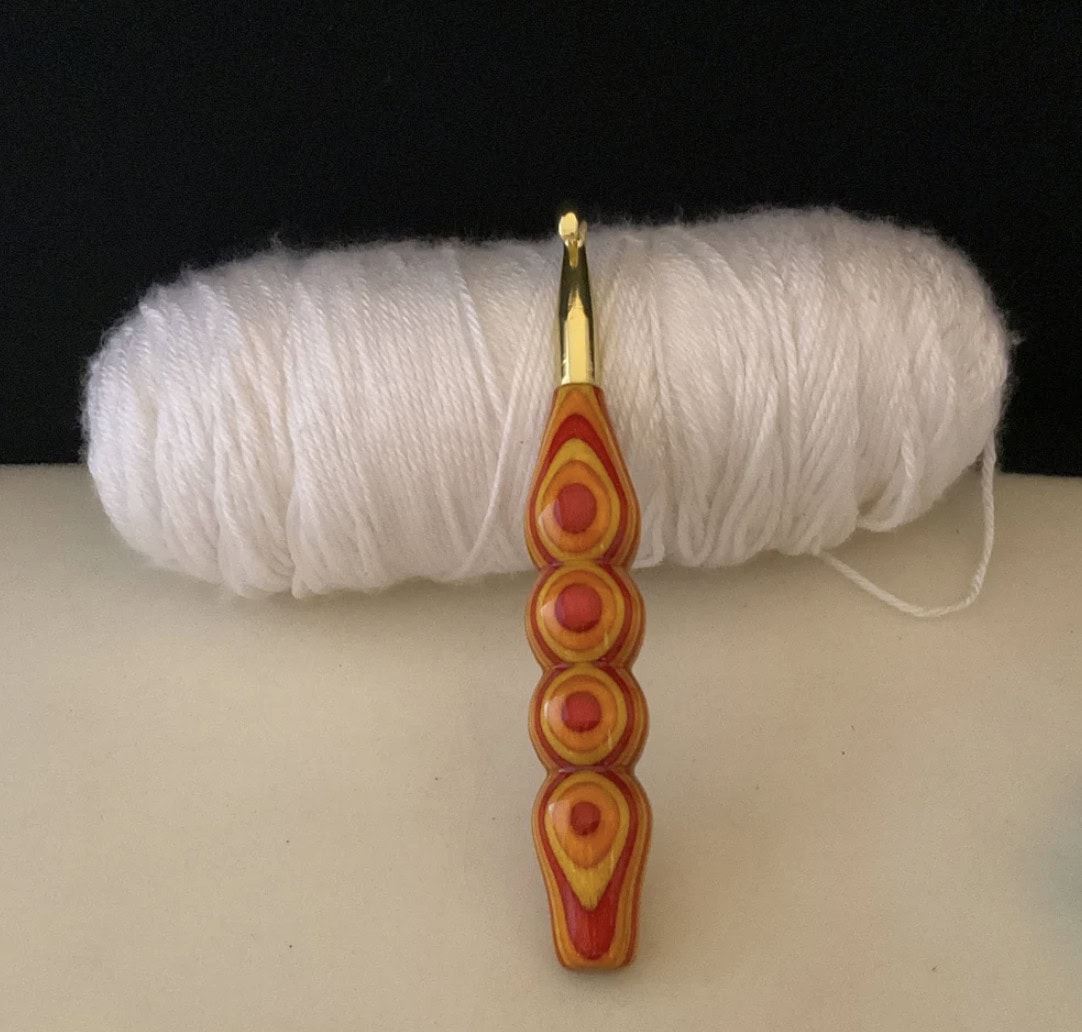 A crochet hook with a colorful, patterned handle stands before a large ball of white yarn, set against a plain background. Nearby, the cheerful Rori the Raptor crochet pattern invites creativity and fun into your crafting journey. -Marly Bird